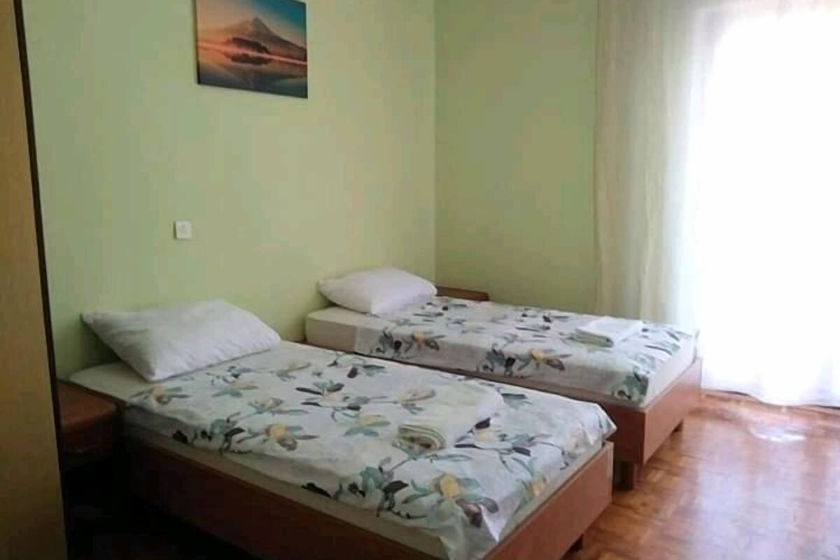 Una habitación con dos camas individuales, cada una con ropa de cama floral y una almohada. Hay un cuadro en la pared sobre las camas y una ventana con cortinas al lado derecho.
