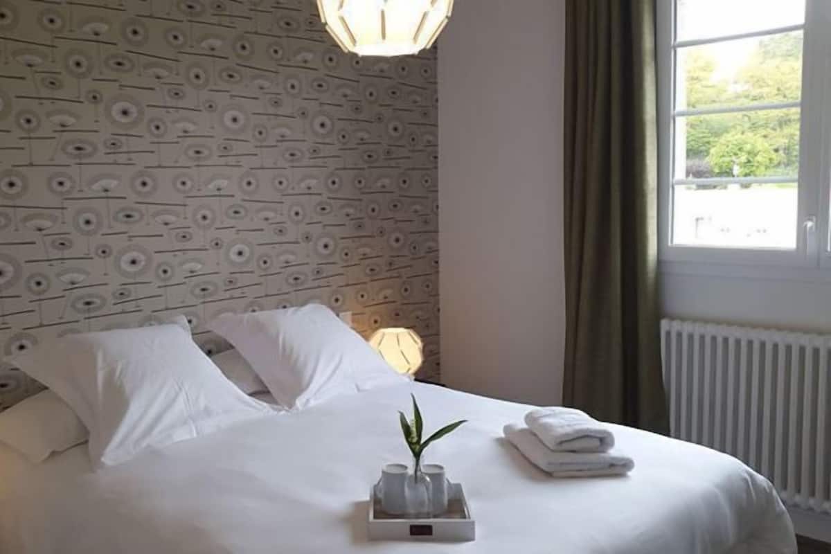 A hotel room with a patterned wallpaper, a neatly made bed with white linens, a bedside lamp, and a window with curtains.