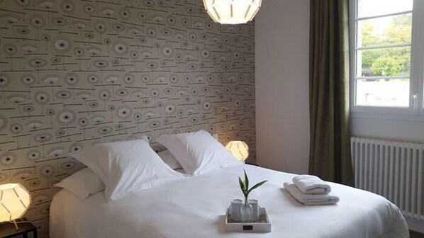Une chambre d’hôtel avec un papier peint à motifs, un lit bien fait avec du linge de lit blanc, une lampe de chevet et une fenêtre avec des rideaux.