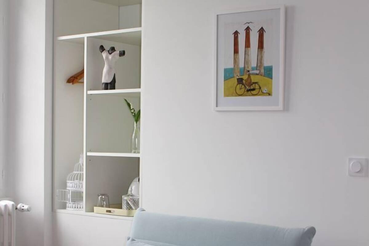 A light blue armchair with a cushion, a white shelving unit with decorative items, and a framed picture on the wall.