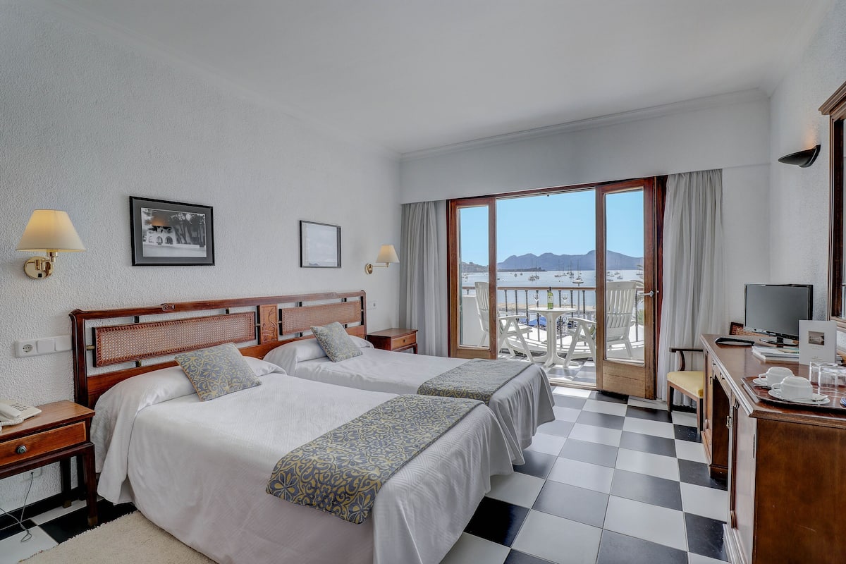 A hotel room with two beds, a desk, a TV, and a balcony with a view of boats and mountains.