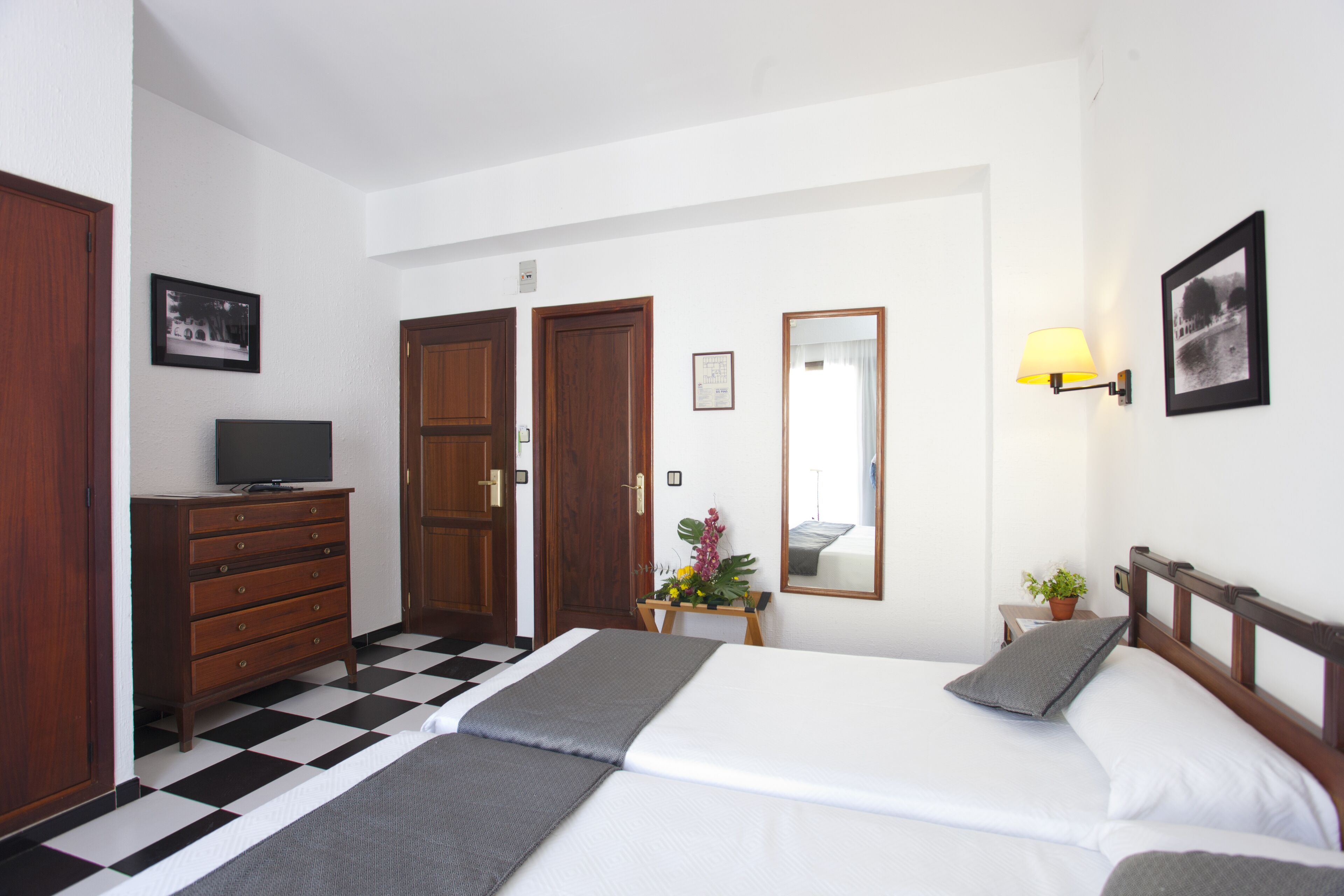 A hotel room with a checkered floor, a bed with white linens, a wooden dresser, a television, a mirror, a nightstand with a lamp, and a framed picture on the wall.