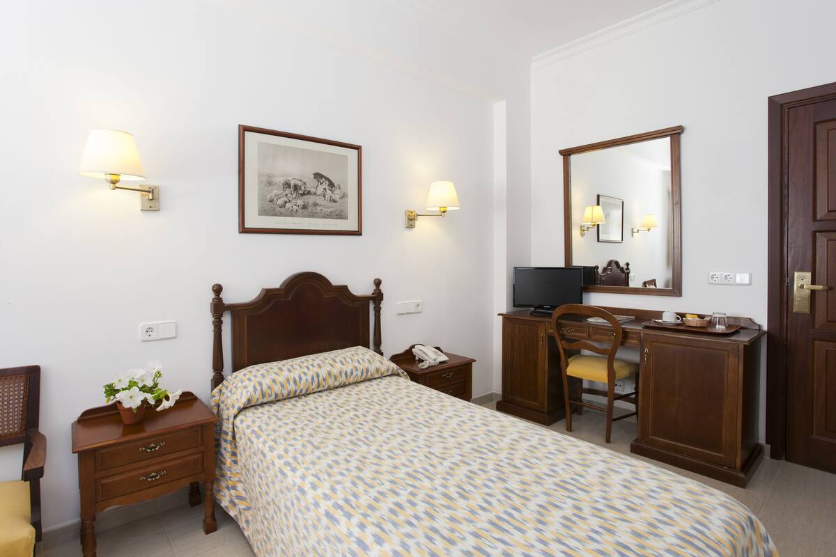 A neatly arranged hotel room with a bed, bedside table, lamp, desk, chair, and a large mirror.