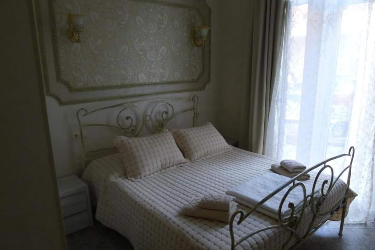 A bedroom with a bed, a nightstand, a wall-mounted light fixture, and a window with curtains.