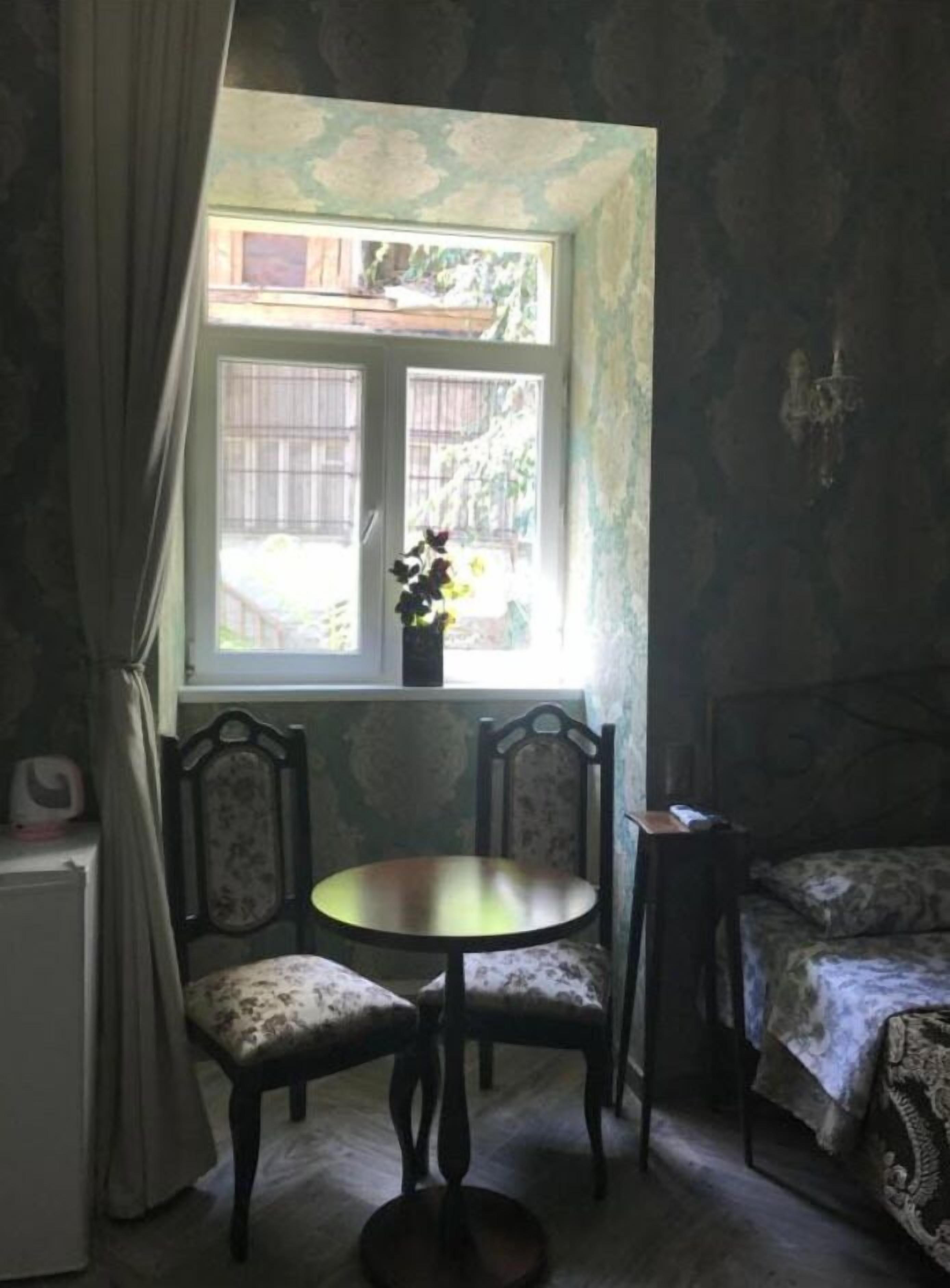A room with a small round table and two chairs, a window with curtains, and a bed with a patterned bedspread.