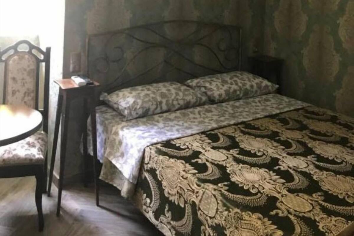 A bedroom with a patterned wallpaper, a bed with a patterned bedspread, a chair, and a chandelier.