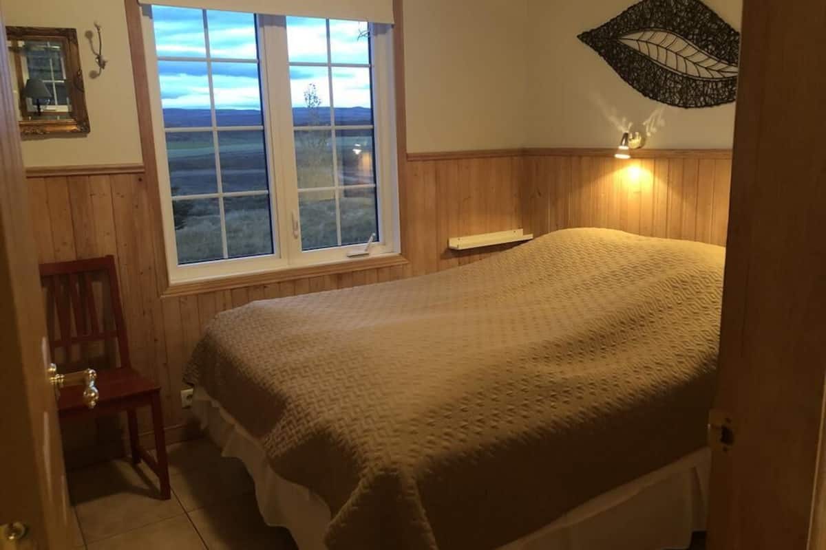 A bedroom with a large bed, a window with a view of the countryside, a wooden chair, and a wall-mounted light fixture.