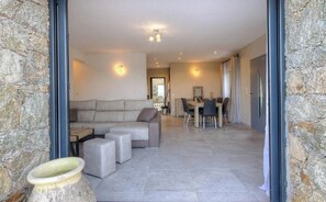 A spacious living area with a sofa, ottomans, and a dining table with chairs. A stone wall and a large window are visible. - Residence Casa Di Roc (Urtaca)