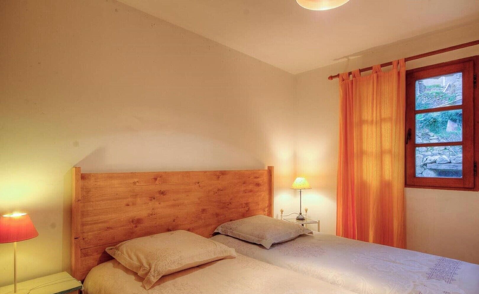 Une chambre à coucher avec une tête de lit en bois, deux lits avec une literie beige, une table de chevet avec une lampe et une fenêtre avec des rideaux.