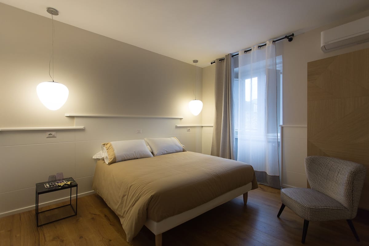 Quarto com cama, mesas de cabeceira, uma cadeira e janela com cortinas.