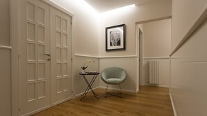 Un couloir doté d’un plancher en bois, une porte blanche ornée d’un motif quadrillé, une petite table avec une lampe, un tableau encadré accroché au mur et une chaise de couleur claire.