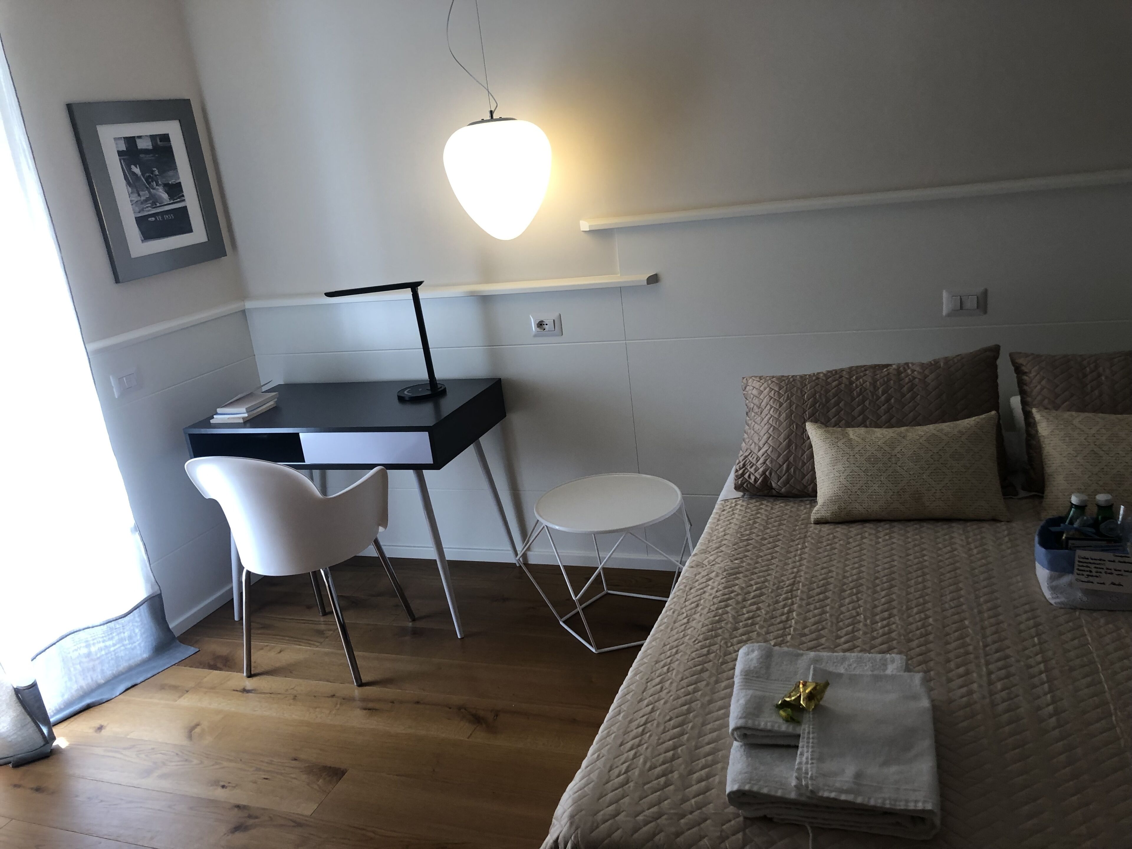 A hotel room with a bed, a desk, a chair, a small table, and a wall-mounted light.