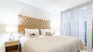 A modern bedroom with a tufted headboard, a large bed with a quilted duvet, two bedside tables with lamps, and a view of a cityscape through sheer curtains. - Waterside Bliss  Austin David Apartments (London)