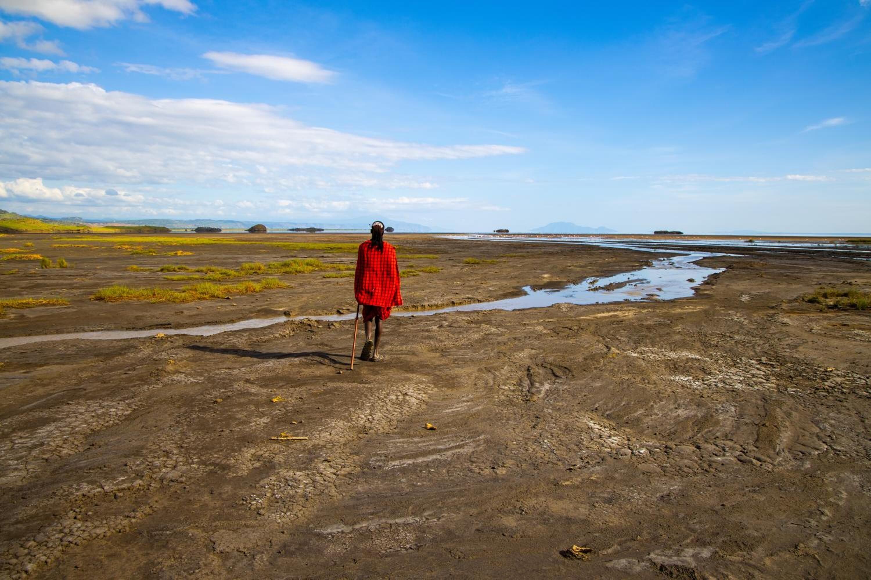 A person in a red garment walking on a muddy terrain with puddles and sparse vegetation.