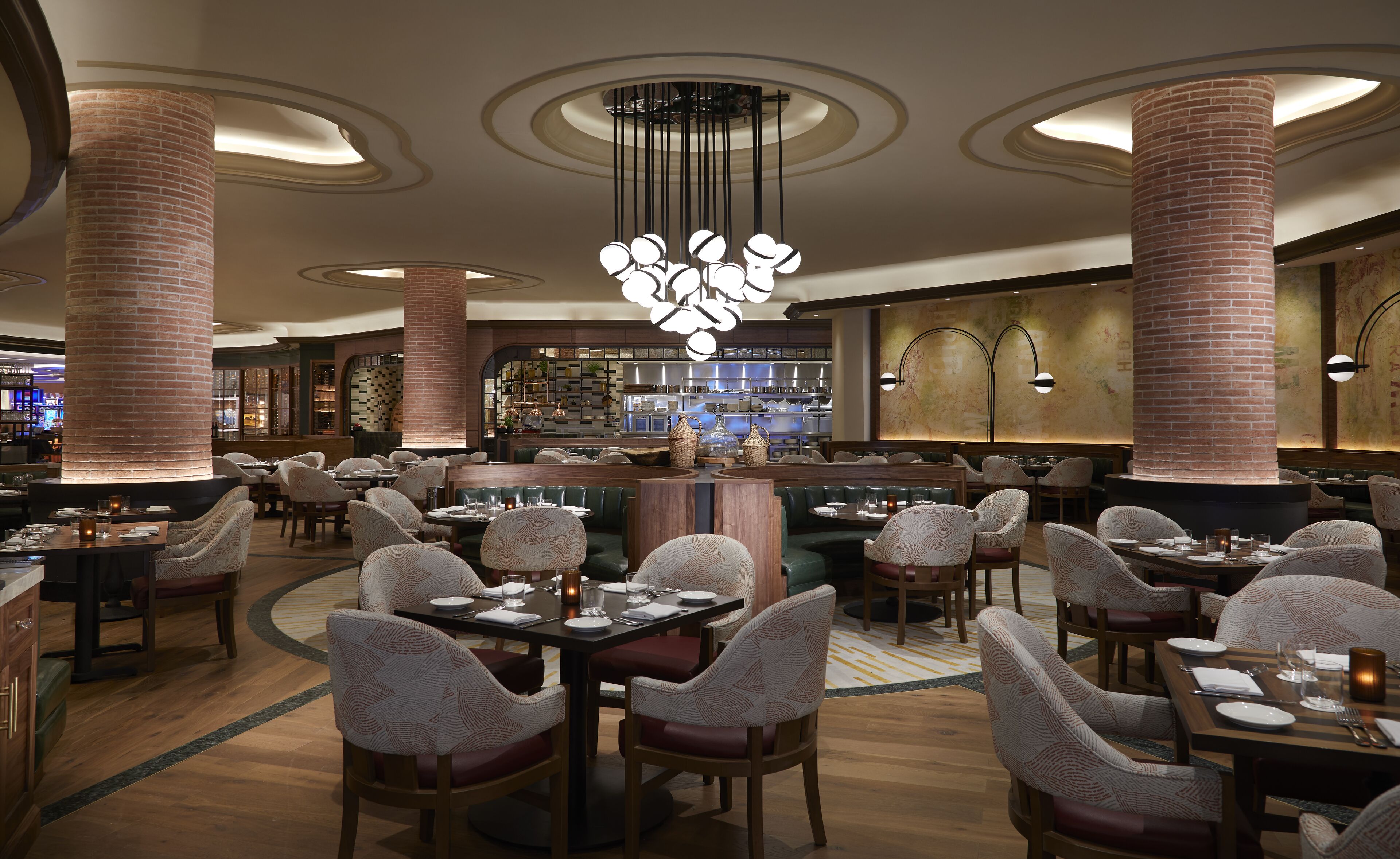 Un restaurant spacieux avec de hauts plafonds, des colonnes en briques apparentes et un grand lustre central. Les tables sont disposées autour d’un bar, avec vue sur la cuisine.