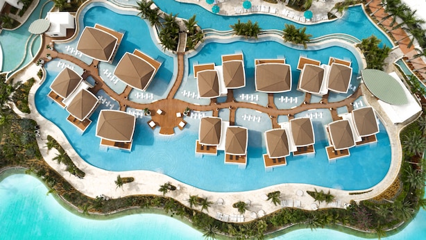 An aerial view of a resort with a large central pool, multiple sun loungers, and surrounding palm trees.