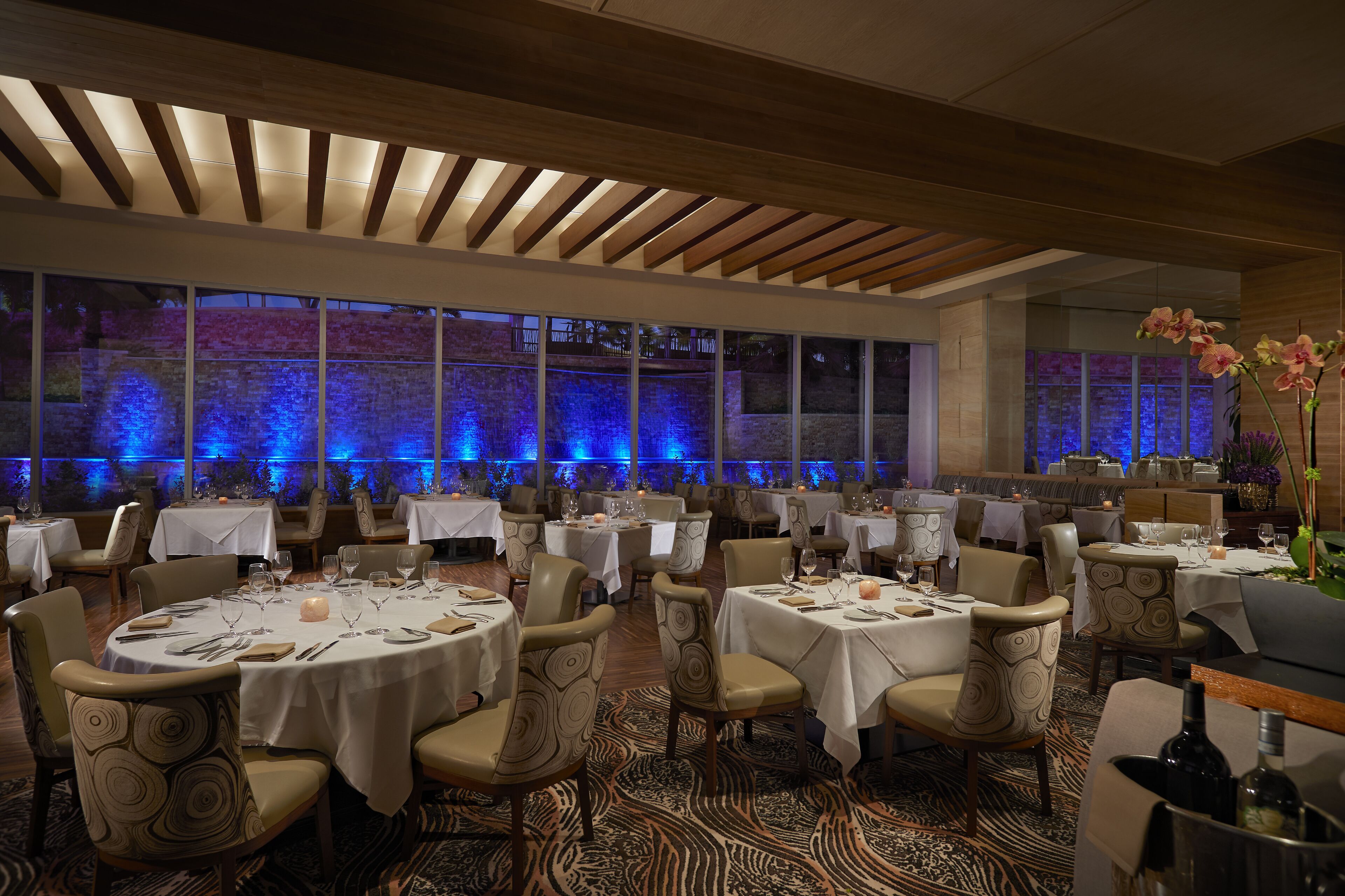 A spacious dining area with round tables and chairs, large windows, and a view of a water feature outside.