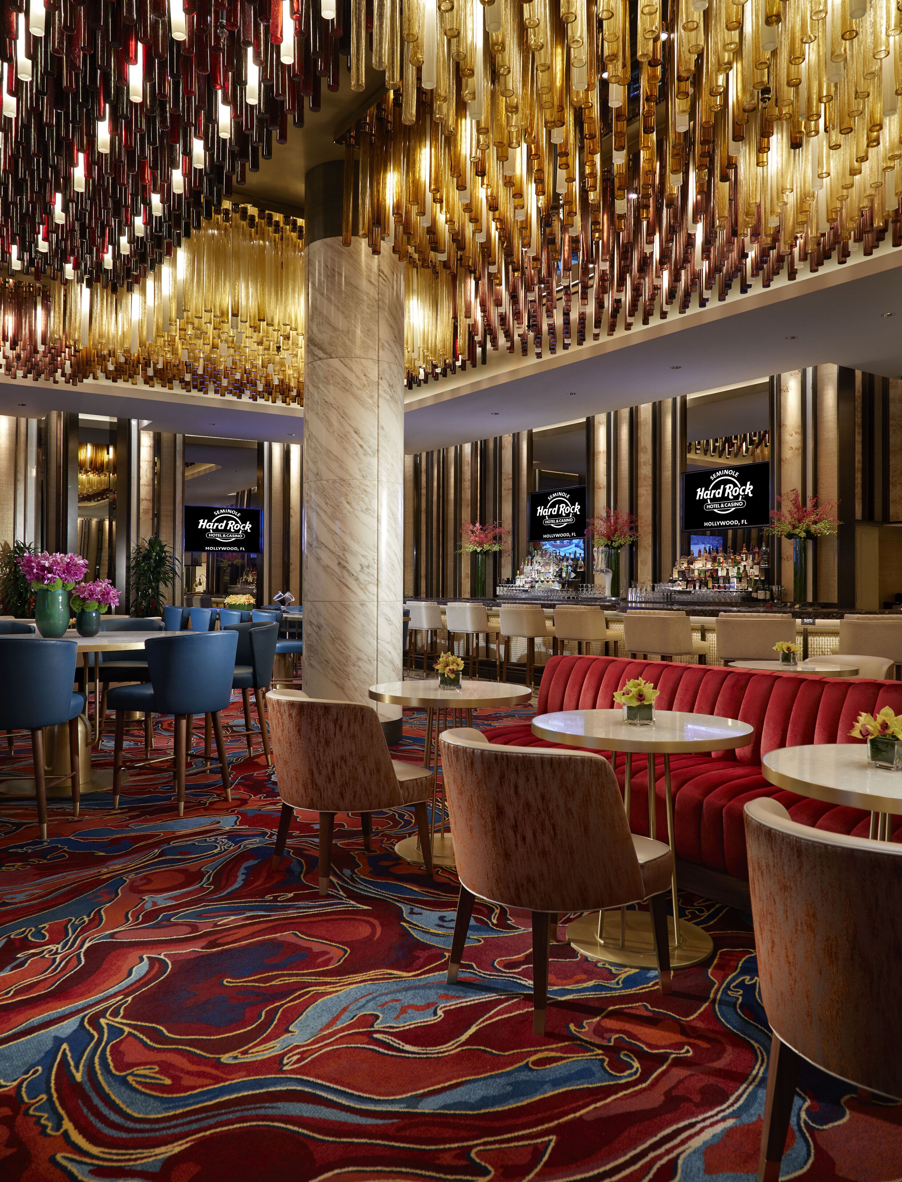 A hotel lobby with a red carpet, round tables, and blue chairs. There is a bar area with a TV screen displaying 'Seminole Hard Rock Hotel & Casino Hollywood, FL'.