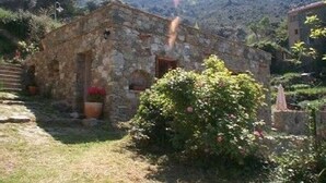 Ein Steinhaus mit einer kleinen Veranda, umgeben von viel Grün und Bergen im Hintergrund.