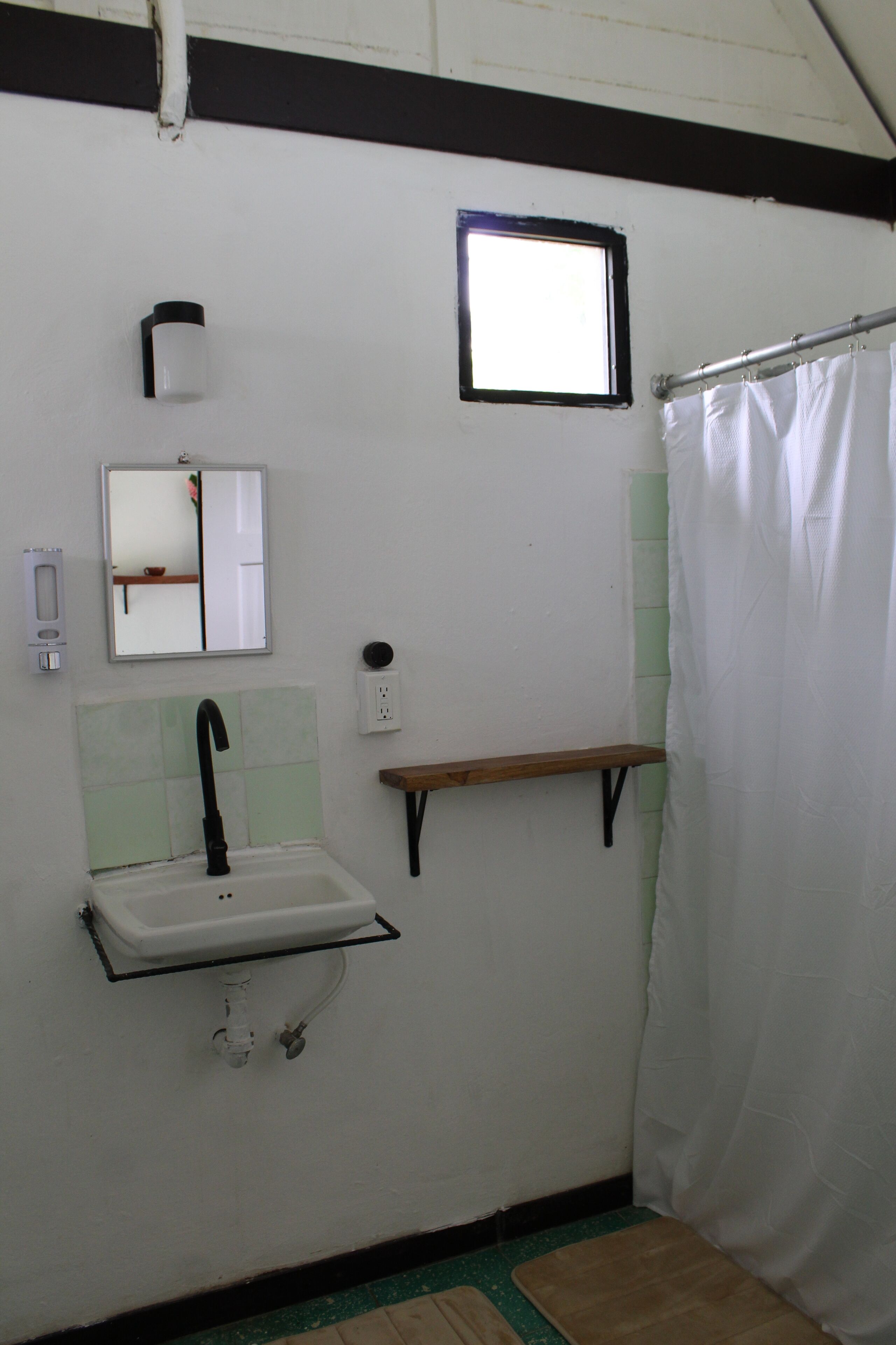 A bathroom with a white sink, a mirror, a window, a shower curtain, and a wooden shelf.
