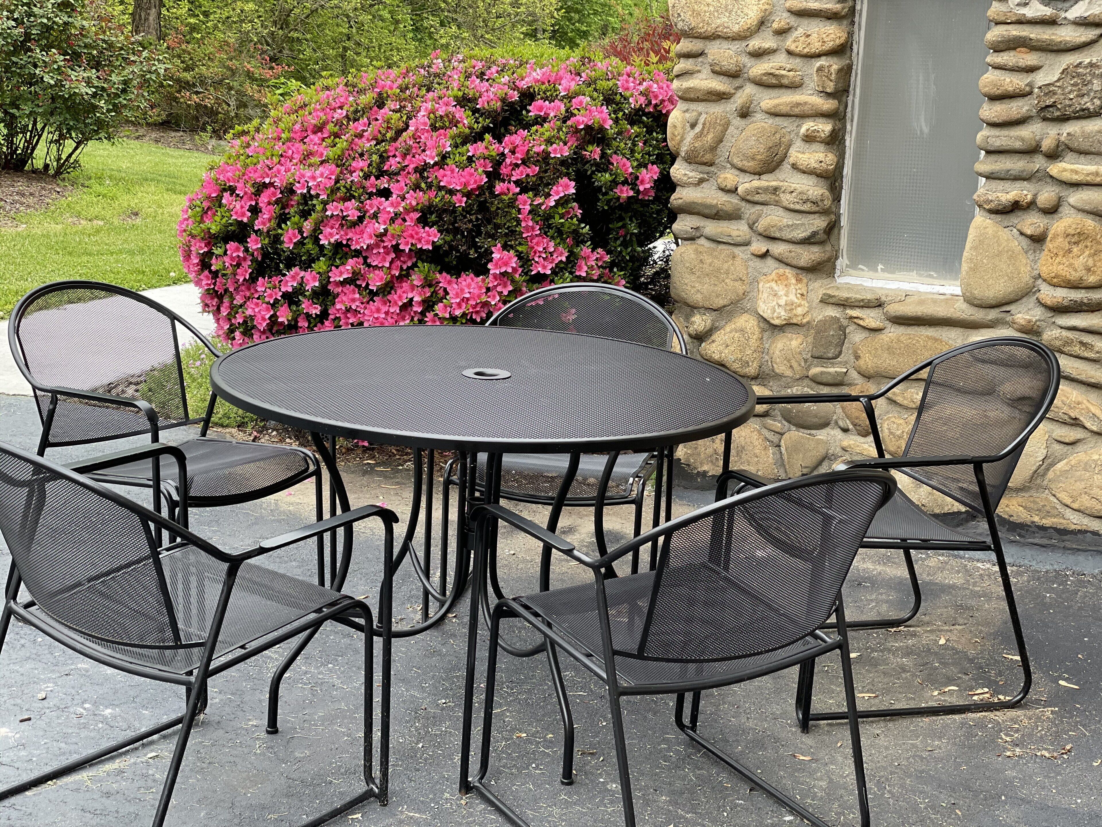 A round outdoor table with six chairs, set against a stone wall and surrounded by pink flowering bushes.