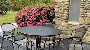 A round outdoor table with six chairs, set against a stone wall and surrounded by pink flowering bushes.