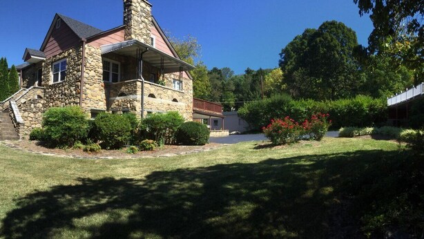 A stone house with a steep roof and a stone chimney, surrounded by well-maintained lawns and trees.