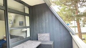 A small balcony with a grey metal table and a grey cushioned chair, a window with white curtains, and a grey wall. - Family apartment in Saint-Lary for 6 people, near the gondola and thermal baths. (Saint-Lary-Soulan)