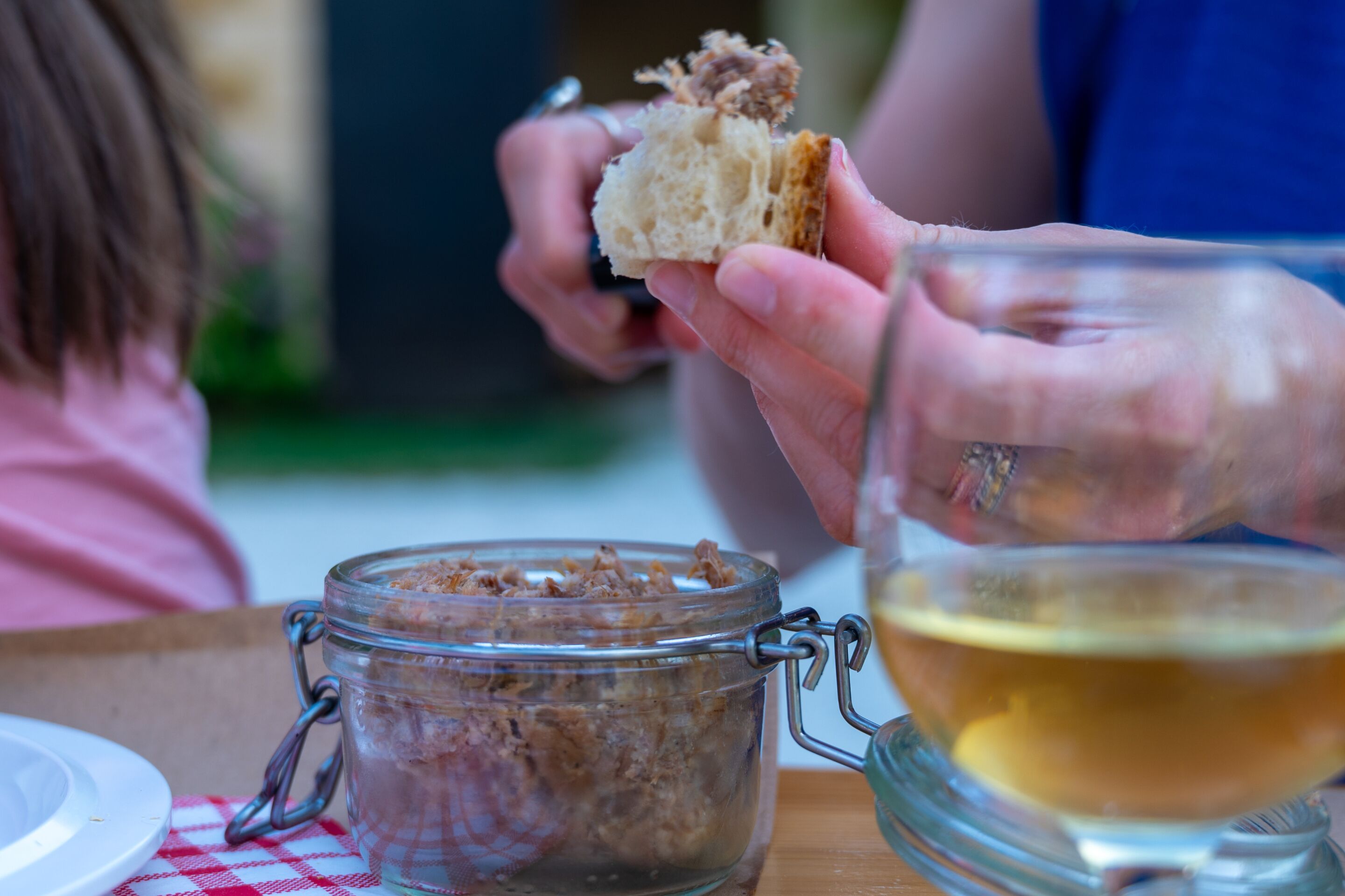 A person holding a piece of bread near a jar of spread and a glass of liquid, possibly tea, on a table with a checkered tablecloth.