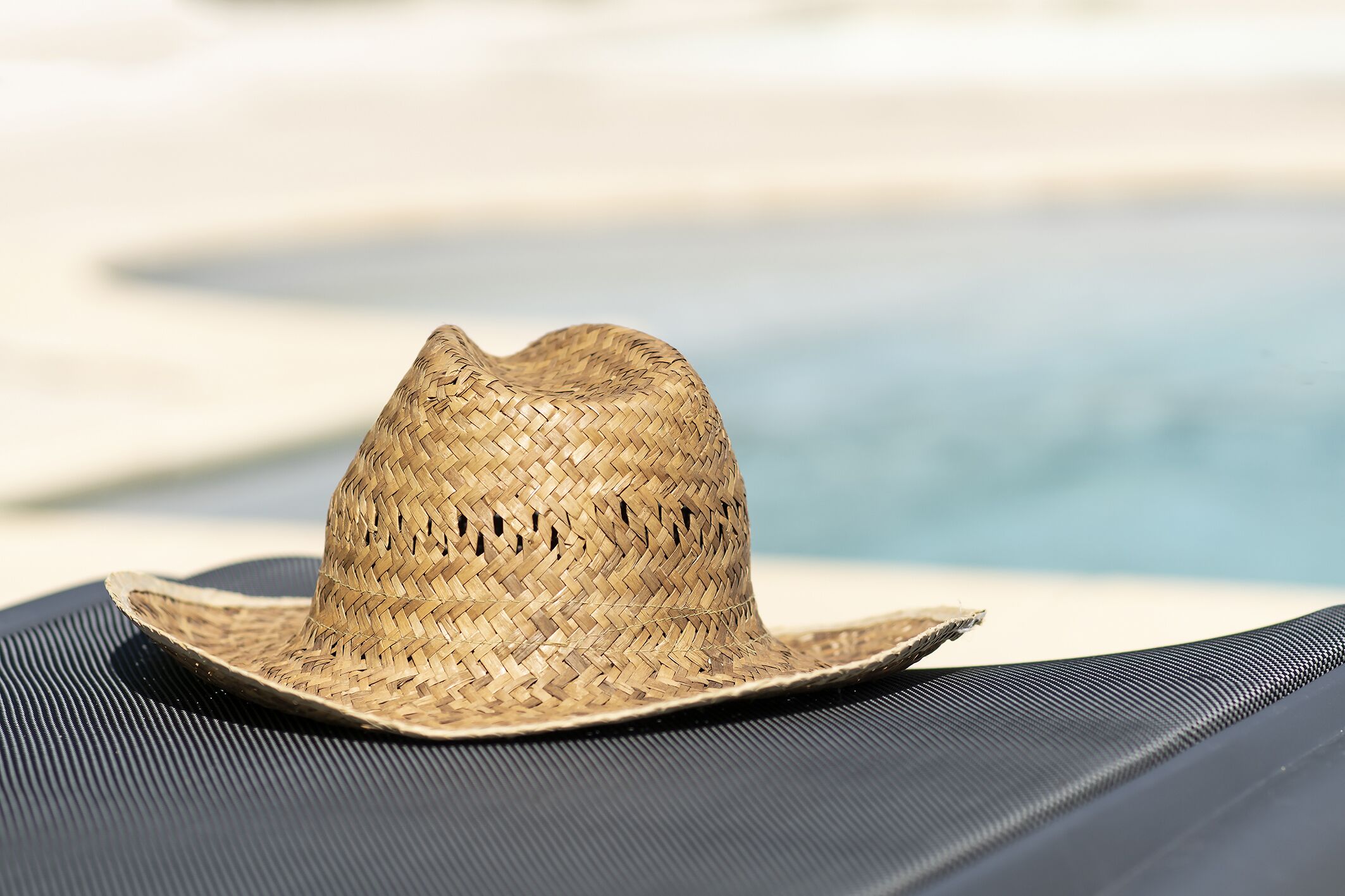 Un sombrero de paja sobre una silla de descanso negra junto a una piscina.