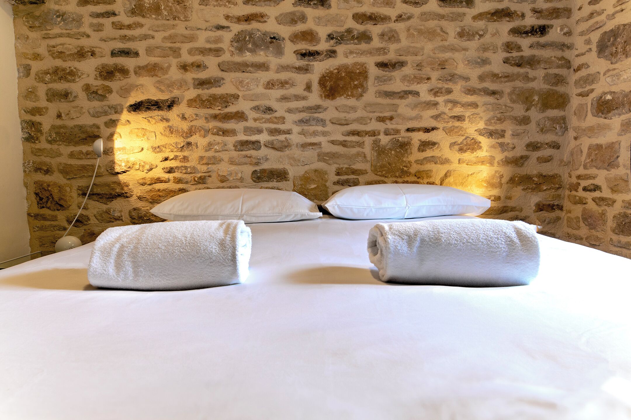 Una habitación con pared de piedra, una cama con sábanas blancas y dos toallas enrolladas sobre la cama.