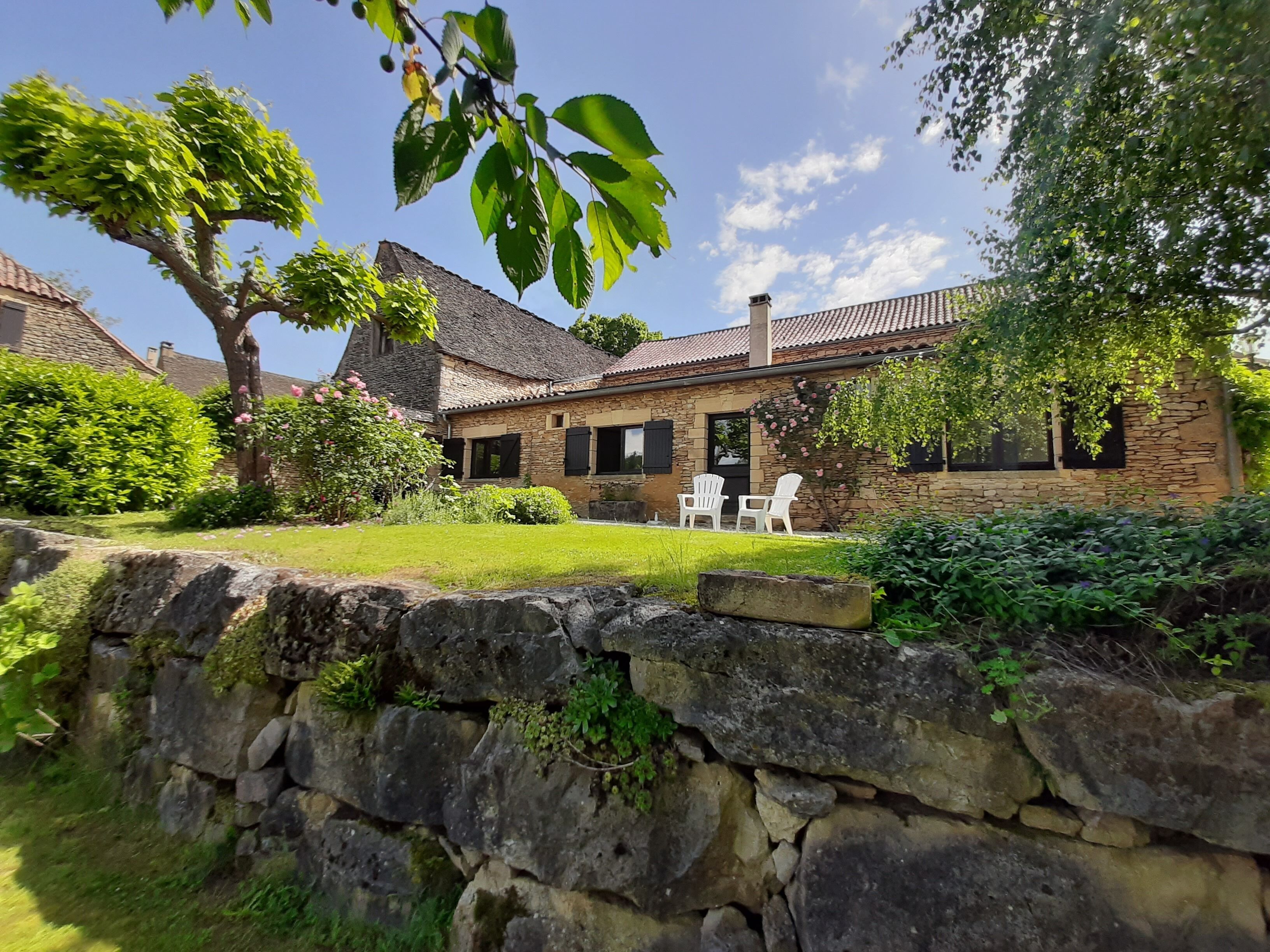 A stone house with a tiled roof, surrounded by a stone wall and lush greenery.