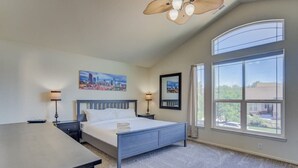 Un dormitorio con cama, mesitas de noche, escritorio, ventanas amplias y obras de arte en las paredes.
