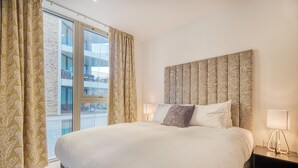 Ein Hotelzimmer mit einem großen Bett, einem Kopfteil, zwei Nachttischlampen und Blick auf ein Gebäude durch ein Fenster.