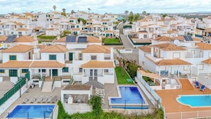 A residential area with multiple white houses, each with a tiled roof and a small front yard. There are two swimming pools visible, one larger and one smaller, surrounded by lounge chairs.