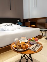 A hotel room with a neatly made bed, a breakfast table with pastries, a bowl of fruit, and a cup of coffee.
