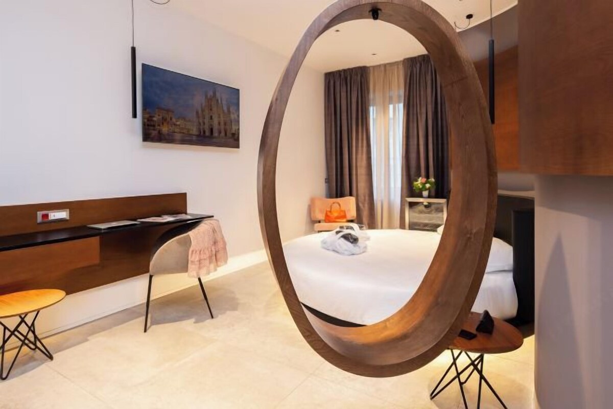 Una habitación de hotel moderna con un espejo ovalado grande, una cama con edredón blanco, un escritorio con una computadora portátil, una silla y una mesita con un bolso de mano.