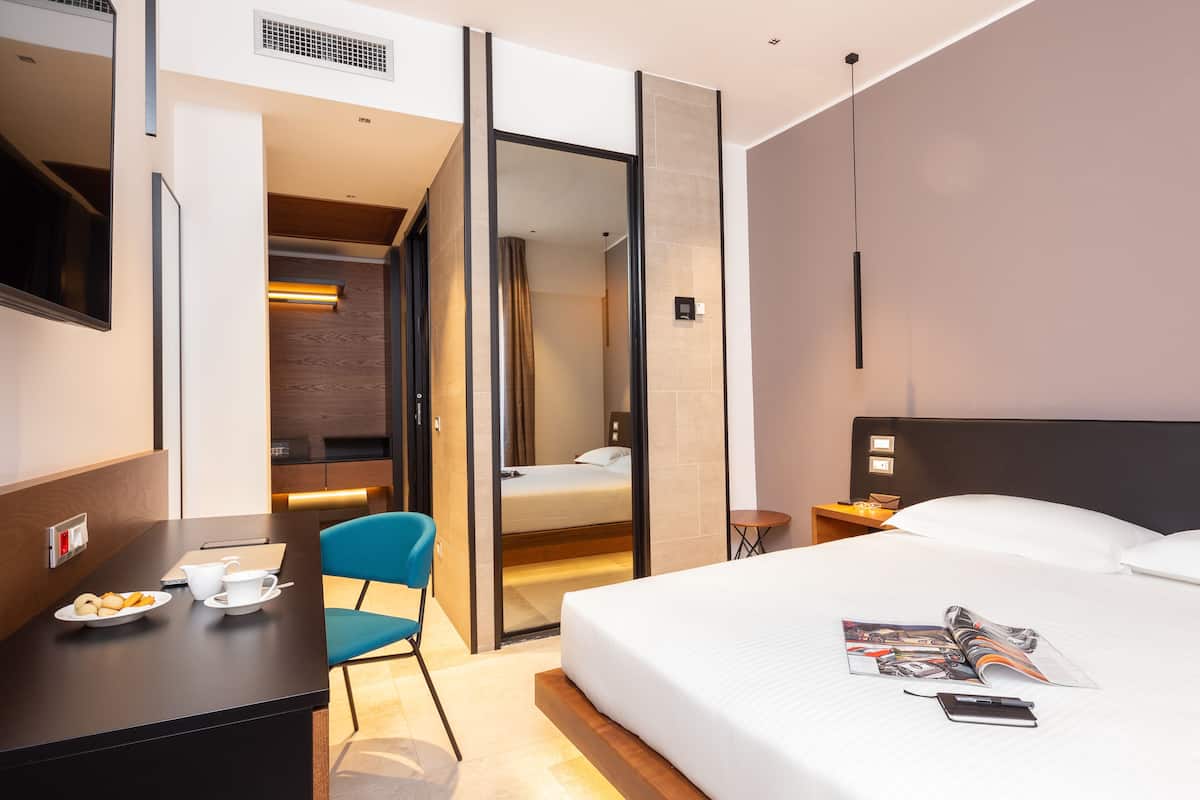 A modern hotel room with a bed, a desk with a chair, a small table, and a TV.