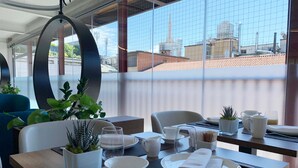 A modern dining area with a table set for two, featuring a city view through large windows. - The Corner Duomo Hotel (Milan)