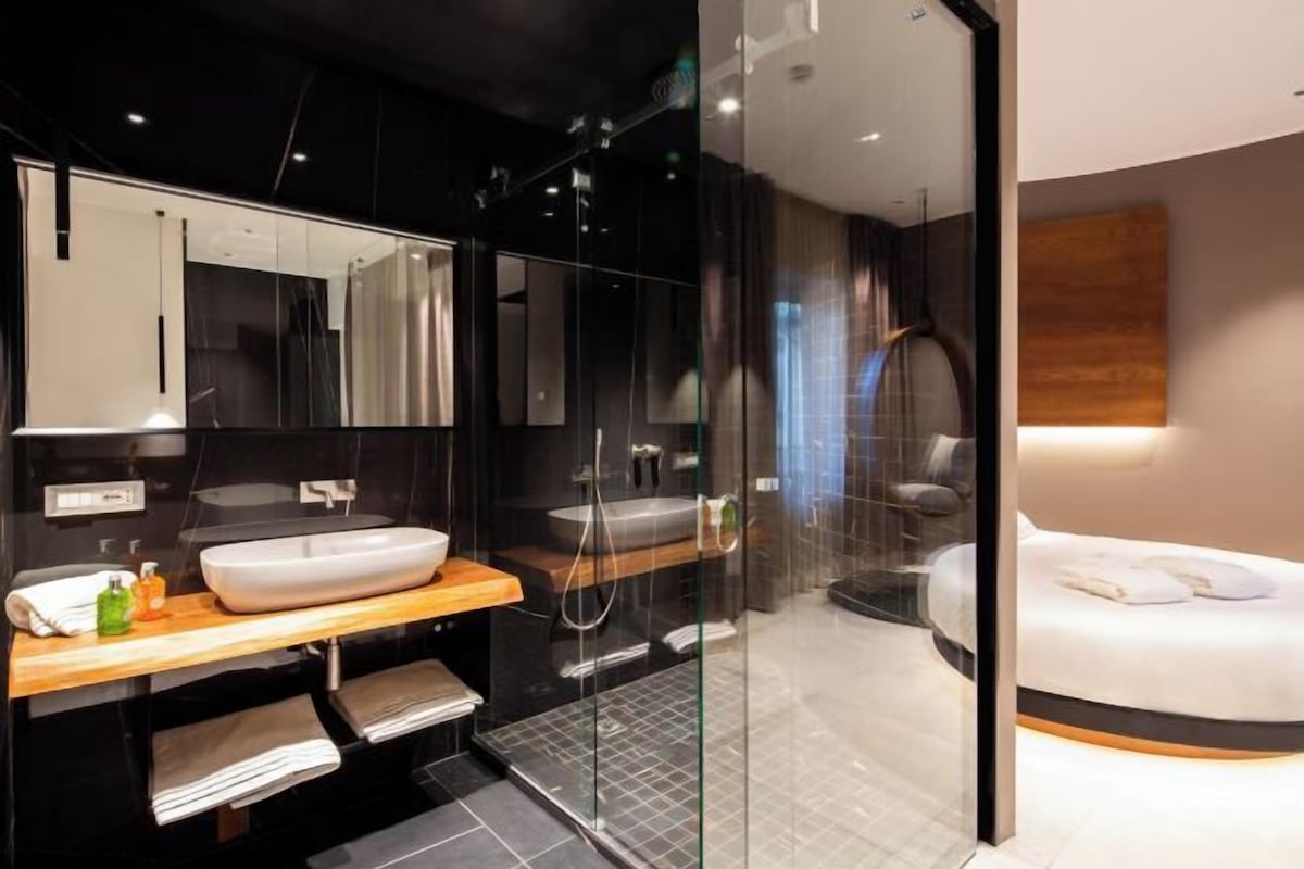 A modern bathroom with a large mirror, two sinks, a shower, and a bed with a wooden headboard.