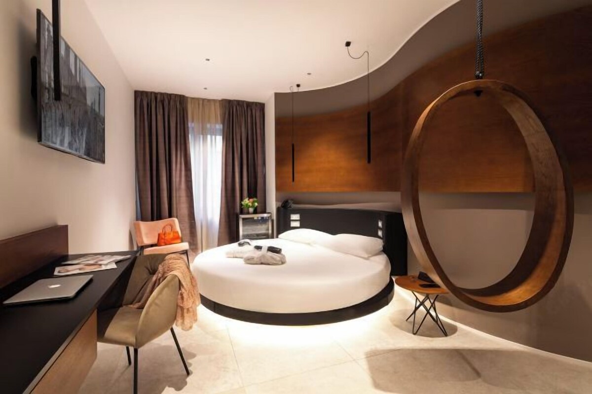 Una habitación de hotel moderna con una cama redonda, un escritorio con una computadora portátil, una silla y una lámpara colgante circular.