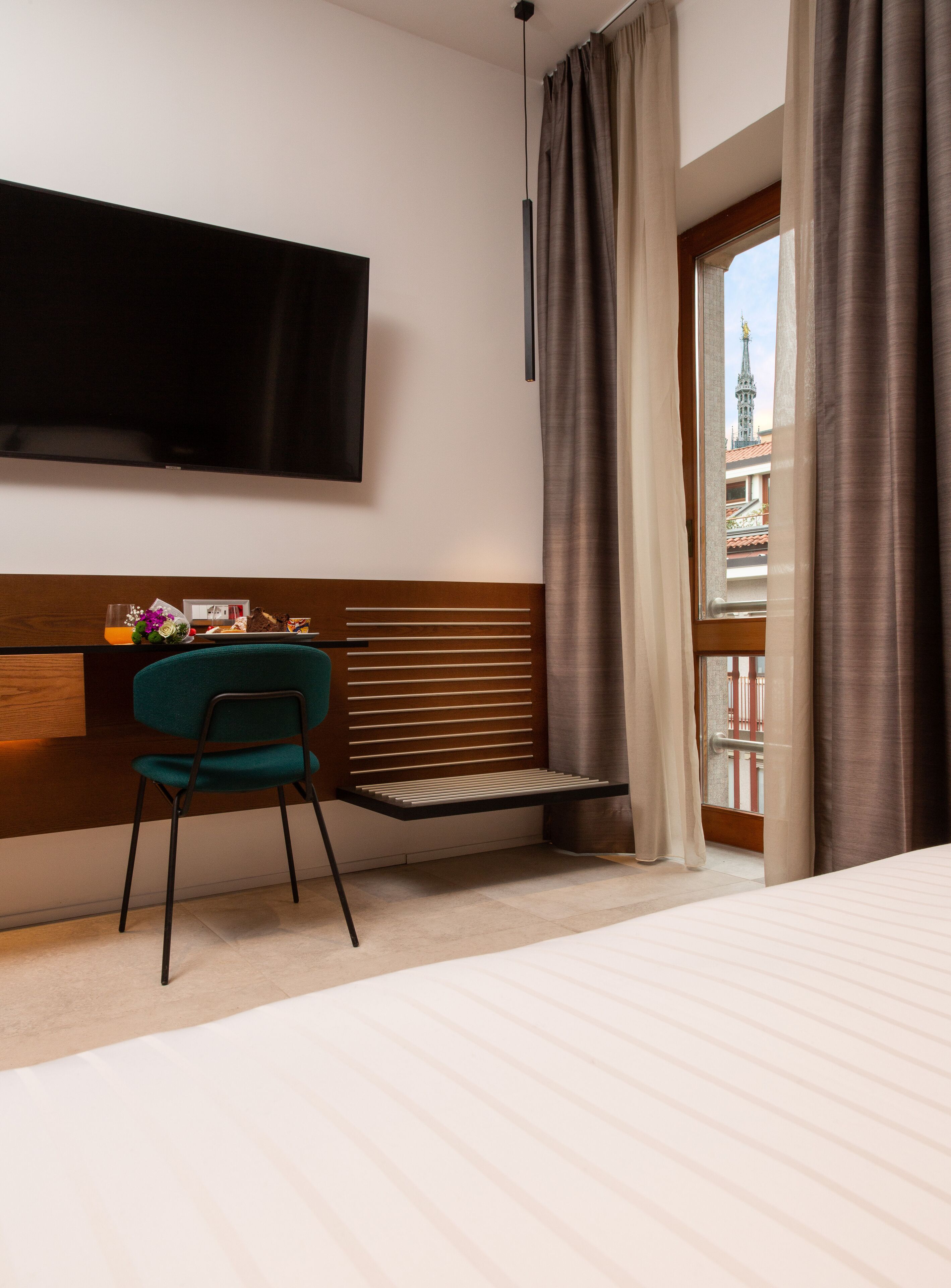 A modern hotel room with a bed, a desk with a chair, a TV mounted on the wall, and a window with curtains.