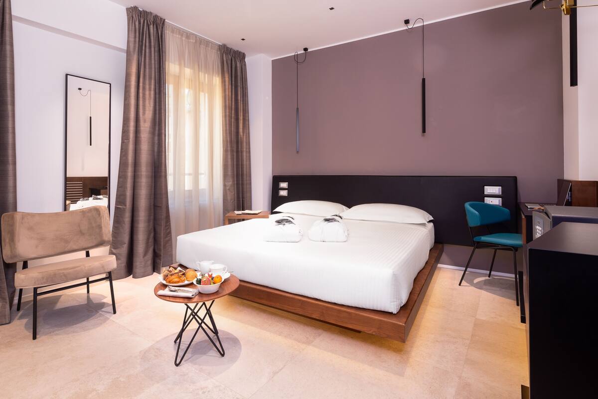 A modern hotel room with a large bed, a desk, a chair, and a small table with breakfast items.