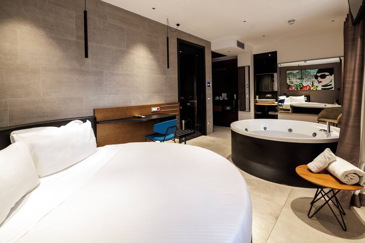 A modern hotel room with a large bed, a round bathtub, a small table with a towel, and a blue chair.