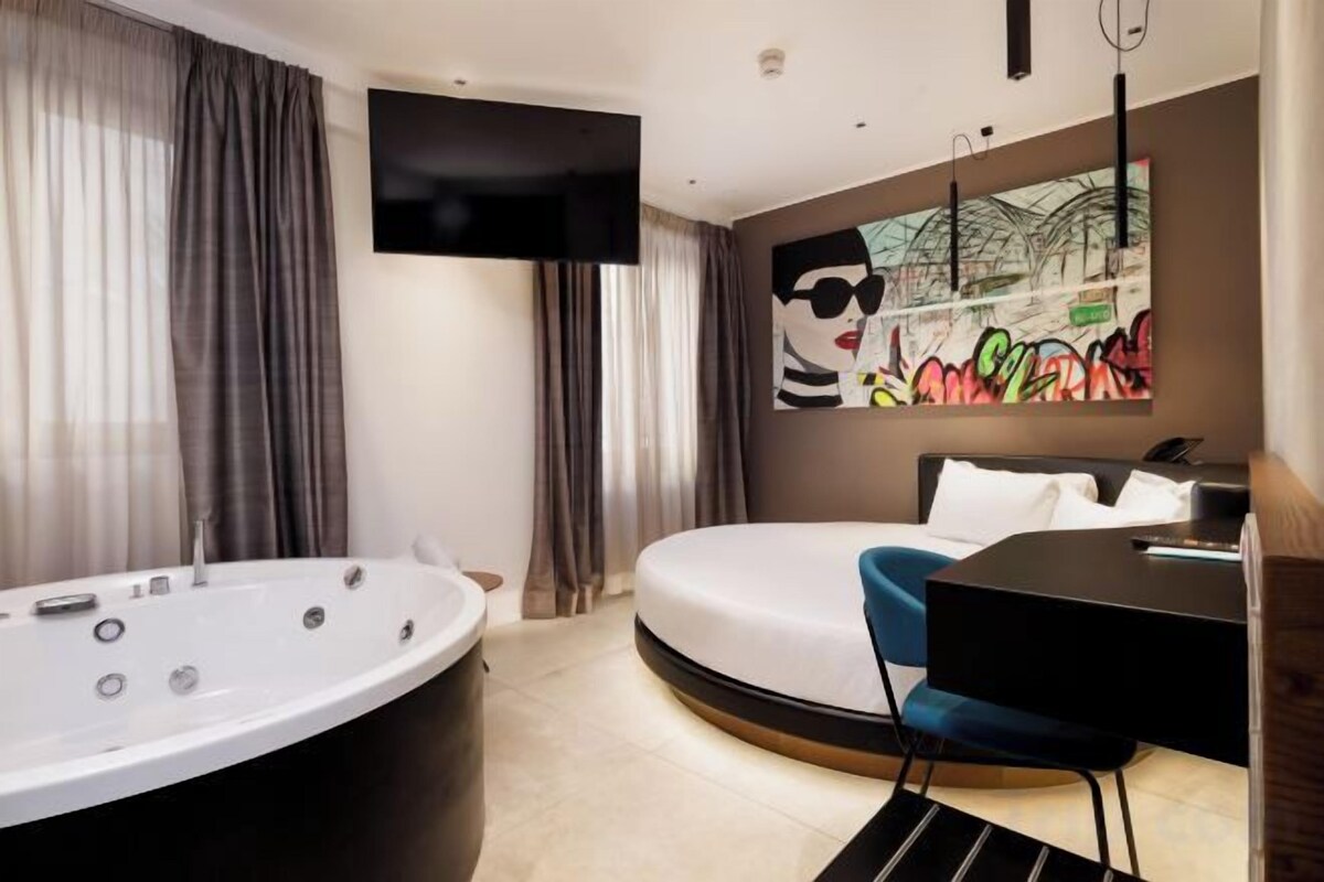 Une chambre d’hôtel moderne dotée d’une grande baignoire, d’un lit rond, d’un bureau et d’un téléviseur fixé au mur.