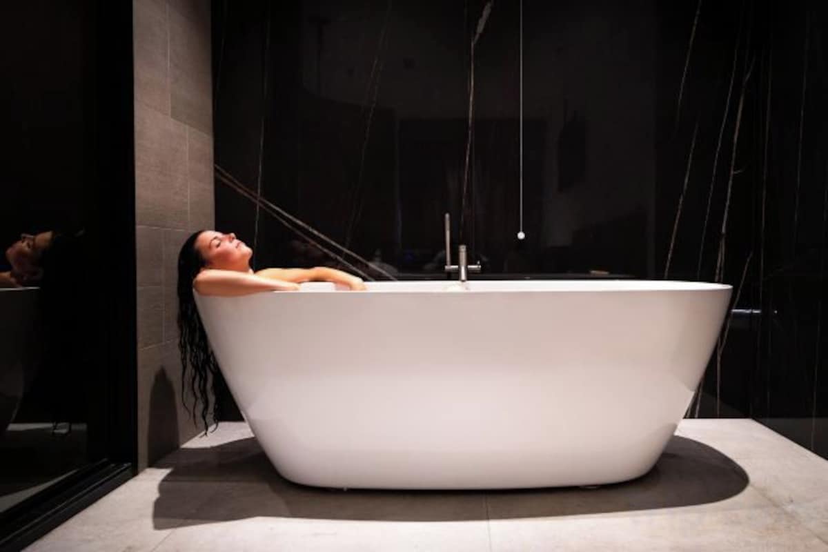 A person relaxing in a modern, freestanding bathtub in a dimly lit room with a dark background.