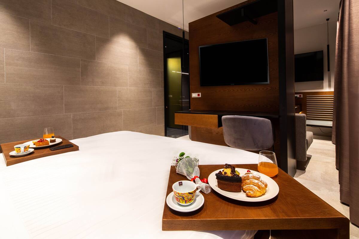 A hotel room with a bed, a wooden side table with breakfast items, a TV mounted on the wall, and a modern bathroom visible through an open door.