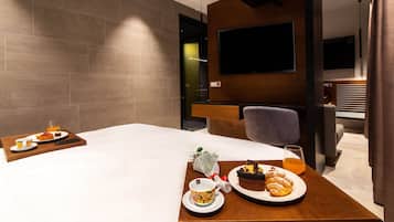 A hotel room with a bed, a wooden side table with breakfast items, a TV mounted on the wall, and a modern bathroom visible through an open door.