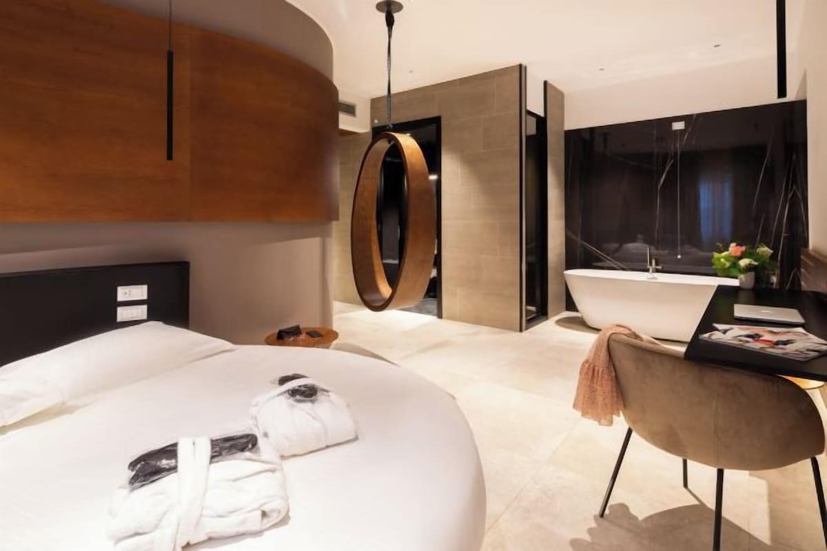 Una habitación de hotel moderna con una cama grande, un espejo redondo, un baño con un espejo amplio y un escritorio con silla.