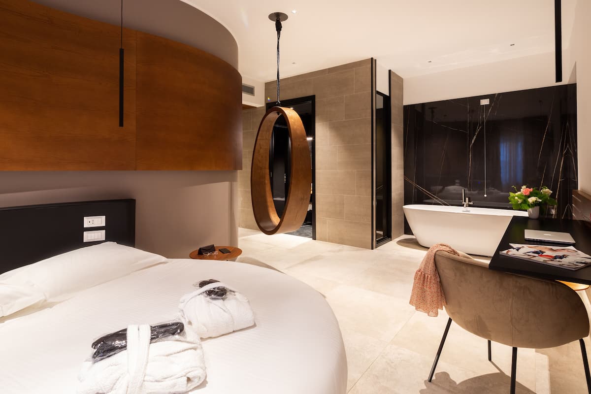 A modern hotel room with a large bed, a round mirror, a bathtub, and a desk with a laptop and magazines.
